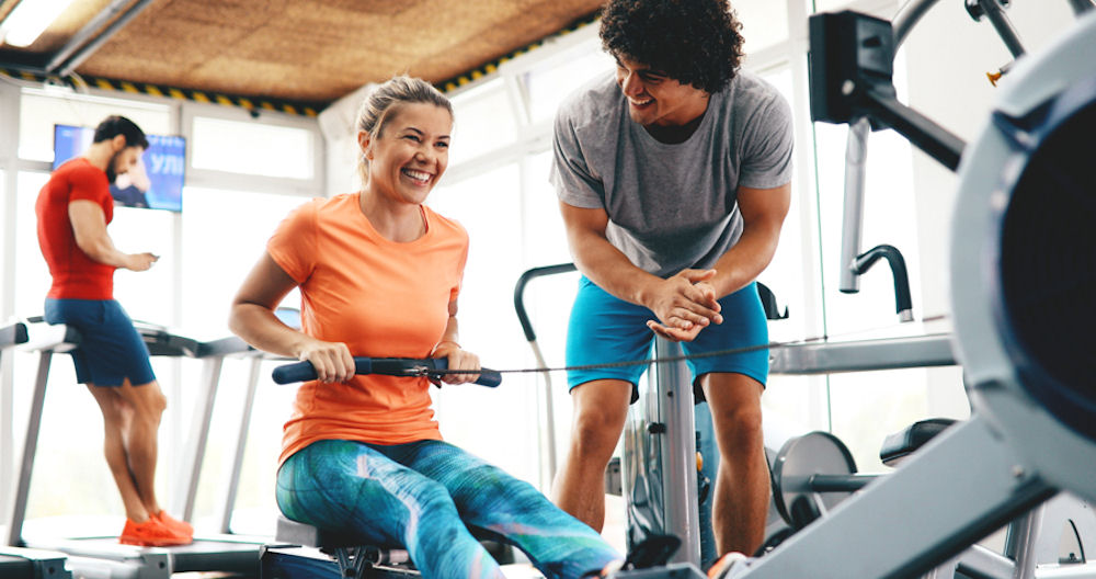 woman on a rowing machine in the gym with a friend to support her