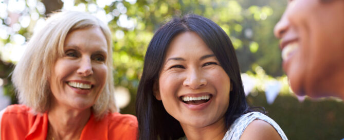Three women sit outdoors, smiling and laughing together. The background is filled with greenery and sunlight, creating a warm and cheerful atmosphere.
