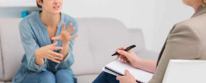 A woman sits on a couch speaking expressively with her hands, while another person faces her, taking notes on a notepad during a conversation or counseling session.