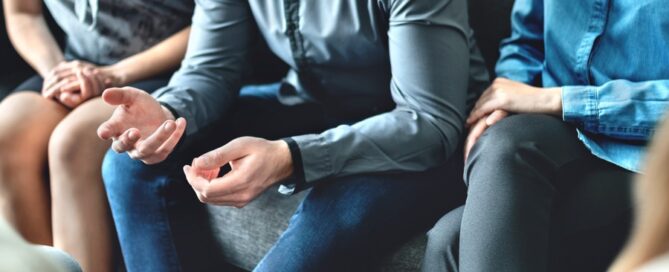 A group of people sit close together on a couch, having a serious conversation. One person gestures with their hands while others listen attentively, suggesting a group therapy session for alcohol addiction.
