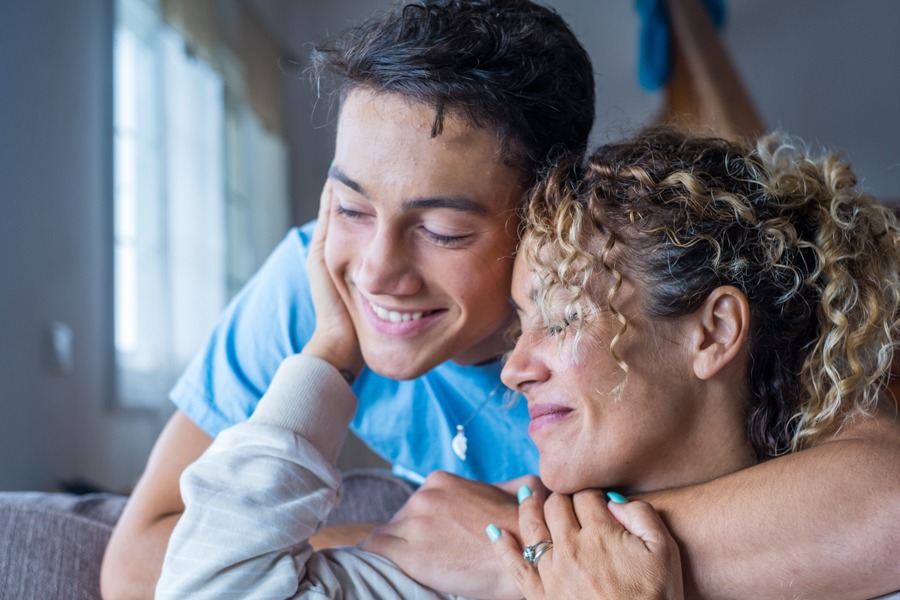 A smiling teenage boy and a woman with curly hair, possibly his mother, share a warm, close embrace indoors—both appearing happy and content, perhaps celebrating progress on their journey through addiction recovery.