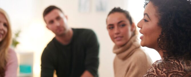 Four people sit in a circle, attentively listening to a woman speak. The informal, supportive setting suggests a group discussion or therapy session focused on drug treatment in a bright, comfortable room.