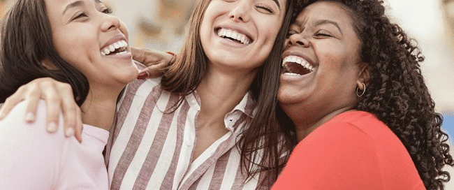 Three women celebrating sobriety stand close together outdoors, smiling and laughing with their arms around each other, enjoying a happy moment. The background is slightly blurred with buildings visible.