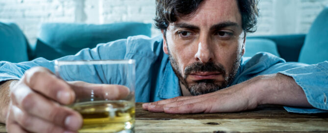 A man with a serious expression rests his arms on a wooden table, staring intently at a glass of whiskey he holds in one hand—perhaps contemplating the impact of alcohol abuse. Blue couch cushions and a white brick wall form the background.
