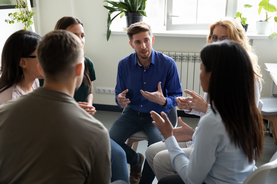 A group of six adults sits in a circle discussing living sober in a bright room with plants on the windowsill. One man in a blue shirt speaks as the others listen and gesture, sharing their journeys to remain substance free.
