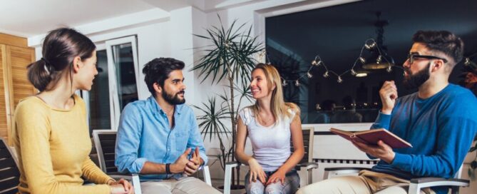 Four adults sit in a circle indoors, engaged in conversation about drug addiction. One person holds an open notebook, while the others listen and smile. The setting is cozy, with modern decor and plants in the background.